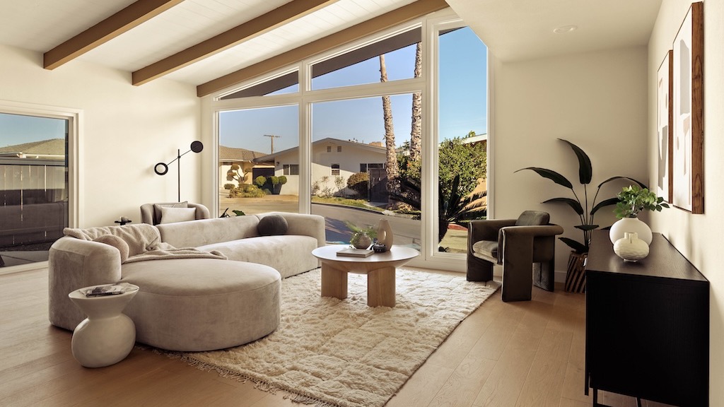 Sunny living room with exposed wood beams, a large window framing palm trees, a curved cream sofa and a round wood coffee table